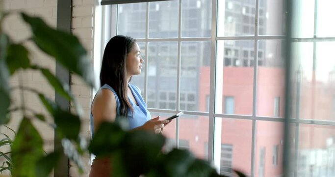 Asian businesswoman stands by a window in an office setting, holding a tablet, with copy space