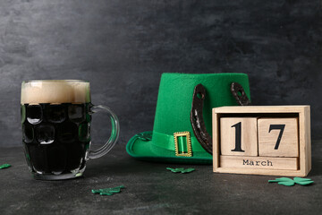 Mug of dark beer with leprechaun hat, horseshoe and calendar on black grunge background. St....