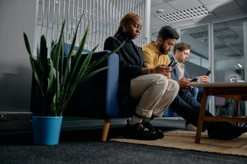 Three multiracial young adults in businesswear sitting on sofa and using mobile phones in office
