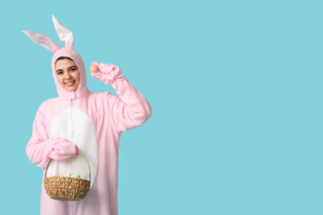 Beautiful young woman in bunny costume holding wicker basket with Easter cake and eggs on blue...