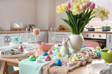Stylish Easter table setting, painted eggs, bunny and vase of flowers in kitchen