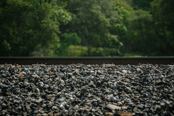 Train tracks are laid on gravel in a rural area. In the foreground, dark gray gravel and the steel railway line cut across the frame. In the background, lush green foliage indicates a forest setting.