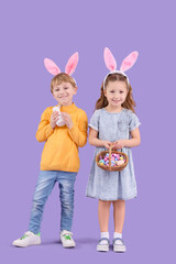 Cute little children in bunny ears holding wicker basket with Easter eggs and rabbit on purple background
