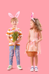 Cute little children in bunny ears holding wicker basket with Easter eggs and bouquet of beautiful tulips on pink background