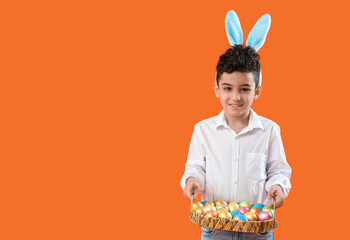 Cute little boy in bunny ears holding wicker tray with Easter eggs on orange background