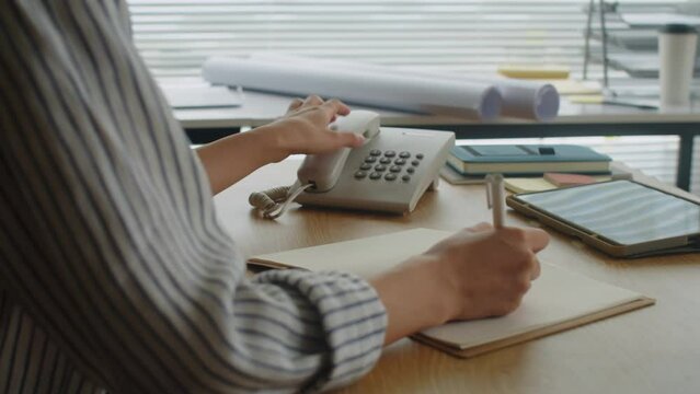 Cropped shot of female manager talking on landline phone with client and making notes sitting at office desk
