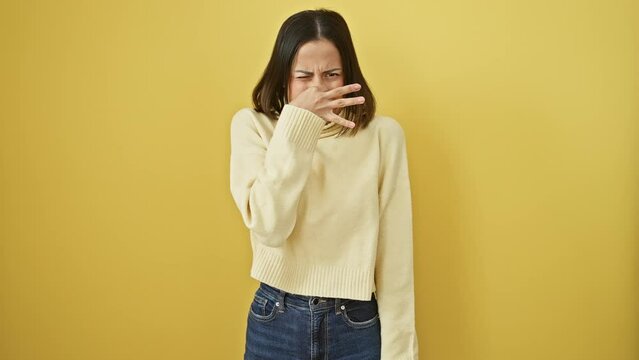 Young beautiful hispanic woman wearing sweater standing smelling something stinky and disgusting, intolerable smell, holding breath with fingers on nose. bad smell over isolated yellow background
