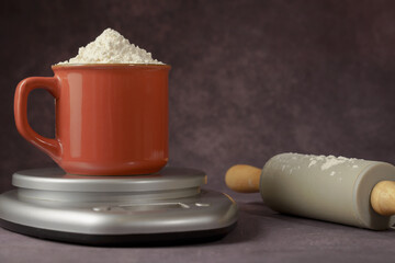 red cup with flour on a kitchen scale next to a rolling pin with a dark background and copy space