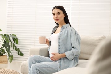 Pregnant woman with cup of drink on sofa at home