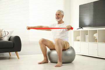 Senior man doing exercise with elastic resistance band on fitness ball at home