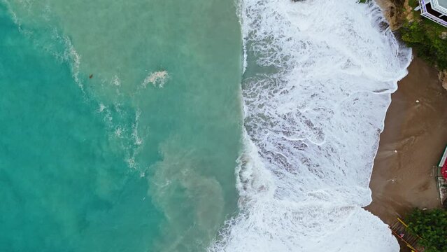 Dark brown sand in cove churns in undertow of strong wave swell in ocean, drone bird's eye view