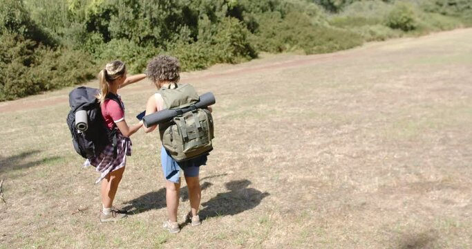 Two women with backpacks are hiking in a sunny, grassy field with copy space