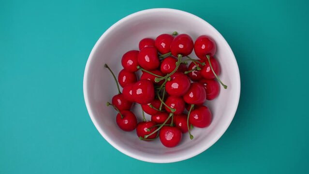 top view of hand putting in red cherries into white bowl isolated blue background