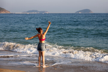 Happy girl in jeans shorts standing on sea beach raising her hands. Freedom, travel and vacation concept