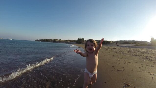 Toddler baby girl in nappy running on sandy beach through sea waves