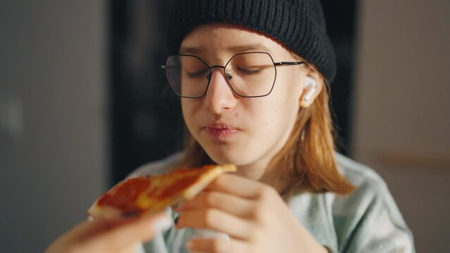 Close up little girl eating pizza food. Hungry young person in glasses and hat is eating piece of pizza, taking a bite from pizza indoor at home. Fast food, children unhealthy tasty food concept.