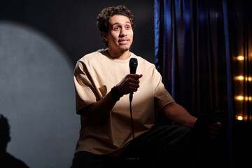 Young male stand up comedian performing his monologue while sitting on stool on stage of concert...