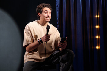 Young comedian with microphone and mobile phone in hands performing monologue in front of audience...