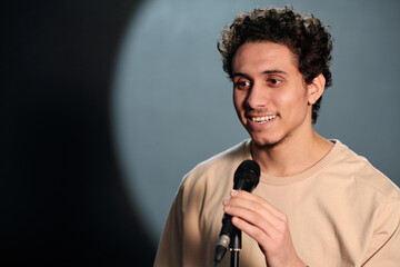 Young smiling man talking in microphone and looking at audience while standing on stage of stand up...