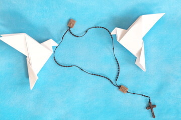 Two white dove origami carrying rosary or scapular in sky blue background.	