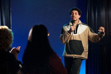 Young comedian in casualwear pronouncing his monologue in microphone while standing on stage and...