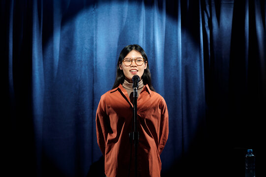 Young Chinese female comedian in brown shirt standing on stage with blue curtains and speaking in microphone during monologue