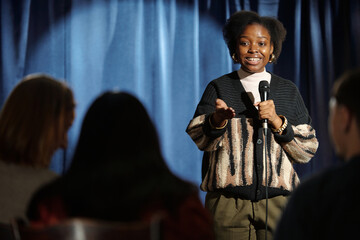 Young African American female comedian standing in front of audience on stage and speaking in...