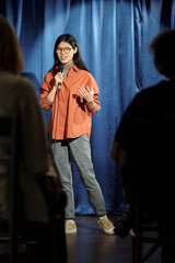 Asian female presenter or participant of stand up show speaking in microphone in front of audience...