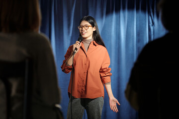 Young female comedian of stand up club standing on stage with blue curtains and pronouncing...