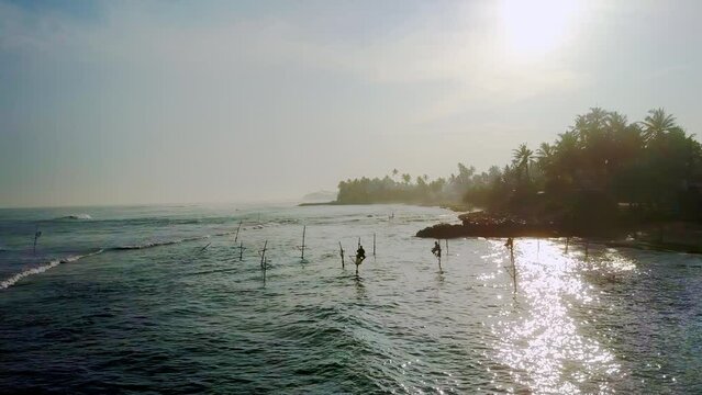 Local fishermen sitting on long wooden poles try to catch fish in wavy ocean lit by sunrise. Exotic island life surrounded by shimmering ocean