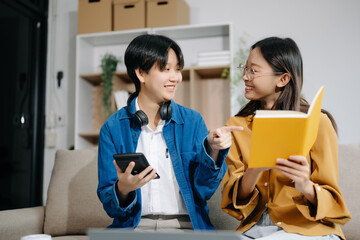 Obraz premium Two Attractive young Asian female college students working on the school project using laptop computer and tablet together, enjoy talking and snacking on sofa
