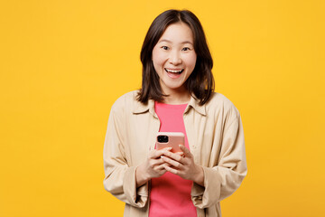 Young surprised shocked woman of Asian ethnicity wear pink t-shirt beige shirt pastel casual clothes hold in hand use mobile cell phone isolated on plain yellow background studio. Lifestyle portrait.