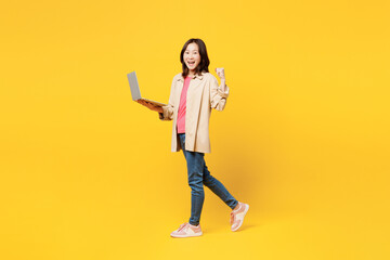 Fototapeta na wymiar Full body young IT woman of Asian ethnicity she wear pink t-shirt beige shirt pastel casual clothes hold use work on laptop pc computer isolated on plain yellow background studio. Lifestyle portrait.