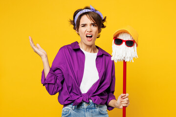 Young confused shocked woman wear purple shirt casual clothes do housework tidy up hold in hand mop in hat and glasses kidding isolated on plain yellow background studio portrait Housekeeping concept - obrazy, fototapety, plakaty