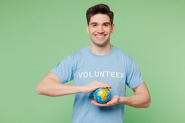 Young smiling man wearing blue t-shirt white title volunteer hold in palms globe look camera isolated on plain pastel light green background. Voluntary free work assistance help charity grace concept.