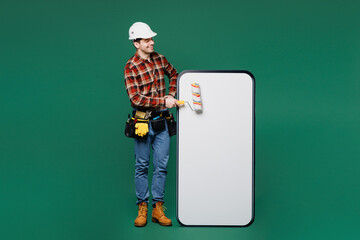 Full body young employee laborer man wear red shirt hardhat paint big huge blank screen area mobile cell phone isolated on plain green background. Instruments for renovation room. Repair home concept.