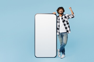 Full body young Indian man wears shirt white t-shirt casual clothes big huge blank screen area mobile cell phone smartphone do winner gesture isolated on plain blue cyan background. Lifestyle concept.