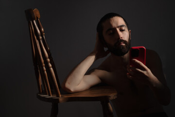 Sitting alone in a dim space, a shirtless man is sitting on the floor, leaning against a chair, lost in thought as he stares at his red cell phone, expressing the loneliness of digital addiction.