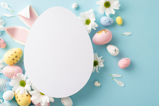 Springtime charm captured in top view photo: pastel-colored eggs, bunny ears, fresh daisies on a soft blue backdrop, complemented by a cut-out card in the shape of an egg for heartfelt Easter messages