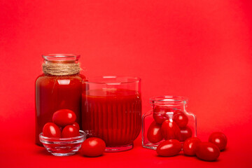 Horizontal photo of a glass of tomato juice and  cherry tomatoes in a square glass jar on a red background.