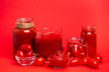 Horizontal photo of a glass of tomato juice, tomato paste and cherry tomatoes in a square glass jar on a red background.