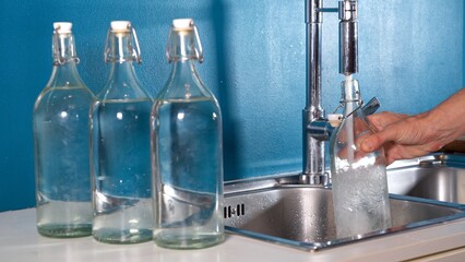 Filling a glass or glass bottle with water from the kitchen tap at home - quality of drinking water in domestic homes. avoid water waste, drought and global warming - obrazy, fototapety, plakaty