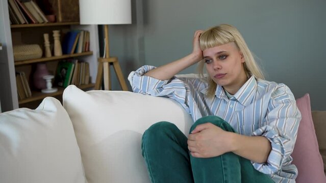 Caucasian sad woman sitting at the sofa with depression. Young student female suffering from mental burnout, feeling depressed, relationship difficulties, sits alone in silence at her home.