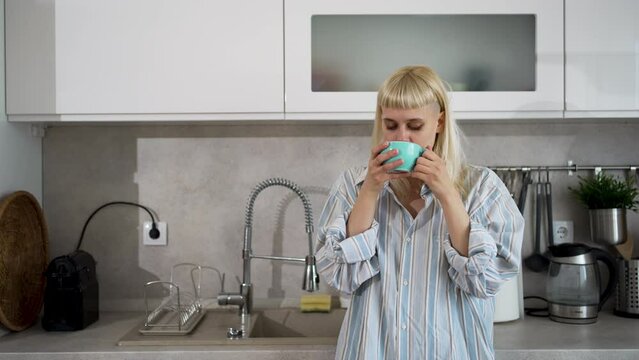 But first coffee. Young happy woman drinking coffee in the kitchen in the morning. Cute blonde female in shirt pajamas enjoying tea as routine in her new apartment home.