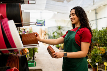 A young Latina woman works at a plant nursery dressed in a dark green apron. The girl is arranging some potted plates on her bookshelf. Concept of working woman.Women in the agronomic sector.