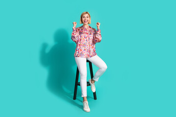 Full body photo of pretty young girl sit chair raise fists winning wear trendy pink print outfit isolated on cyan color background