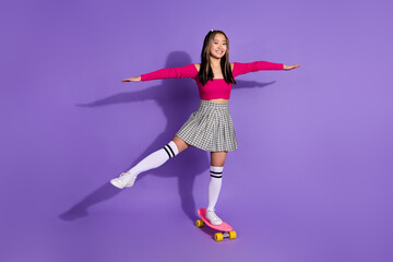 Fototapeta na wymiar Full length photo of positive sweet girl wear pink top arms sides riding skateboard isolated purple color background