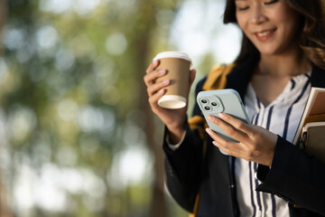 Smiling businesswoman drinking coffee and using mobile phone while walking thought the park