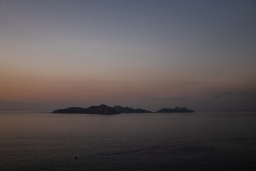 Calm seas at dusk: shot from Monteferro capturing the Atlantic Ocean and Cies Islands' silhouette in the center. Low gray clouds contrast with the orange sky, as a lone boat cruises into twilight.
