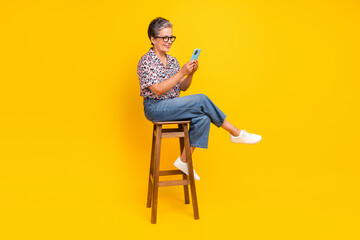 Full body photo of attractive pensioner woman hold gadget sit chair wear trendy leopard print clothes isolated on yellow color background